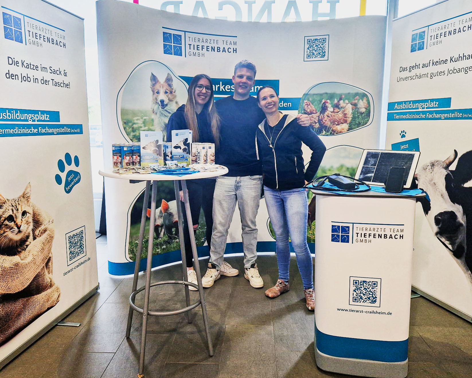 Drei Mitarbeiter*innen des Tier&auml;rzte Teams Tiefenbach stehen an einem Messestand mit Infomaterialien und Tiermotiven &ndash; der Stand informiert &uuml;ber Tiergesundheit, Ausbildung und Praxisangebote.