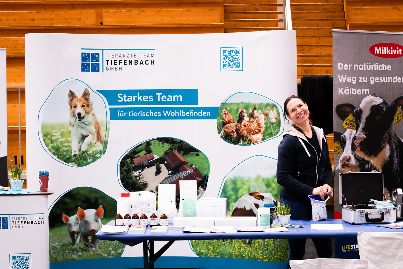 Mitarbeiterin des Tier&auml;rzte Teams Tiefenbach am Messestand mit gro&szlig;em Banner und Informationsmaterialien &ndash; der Stand wirbt mit dem Slogan &sbquo;Starkes Team f&uuml;r tierisches Wohlbefinden&lsquo; und zeigt Bilder von Tieren und der Praxis.